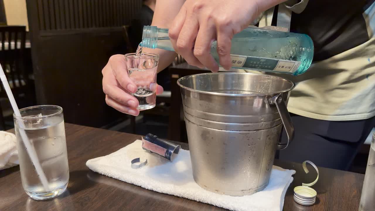 Server pours cold sake from bottle into glass beside ice bucket in warmly lit restaurant