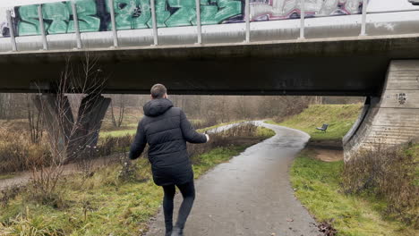 happy man with coat jumping high on rural path with urban bridge in background