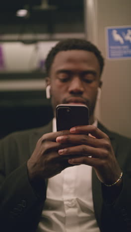 vertical video of young businessman in suit commuting to work sitting in london underground tube train wearing wireless earbuds to stream from mobile phone shot in real time 4