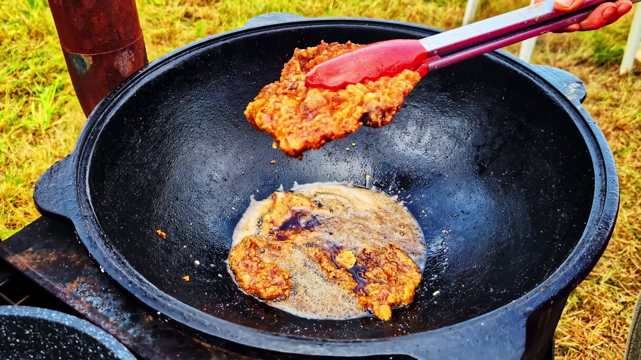 Tongs lift hot fried meat above grill showing crisp crust and fresh oil dripping