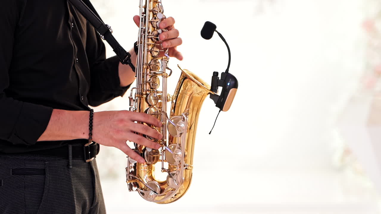 Saxophonist in black shirt and trousers plays jazz on golden saxophone with microphone. Musician male performs music on a saxophone against the blurred background. Live performance.