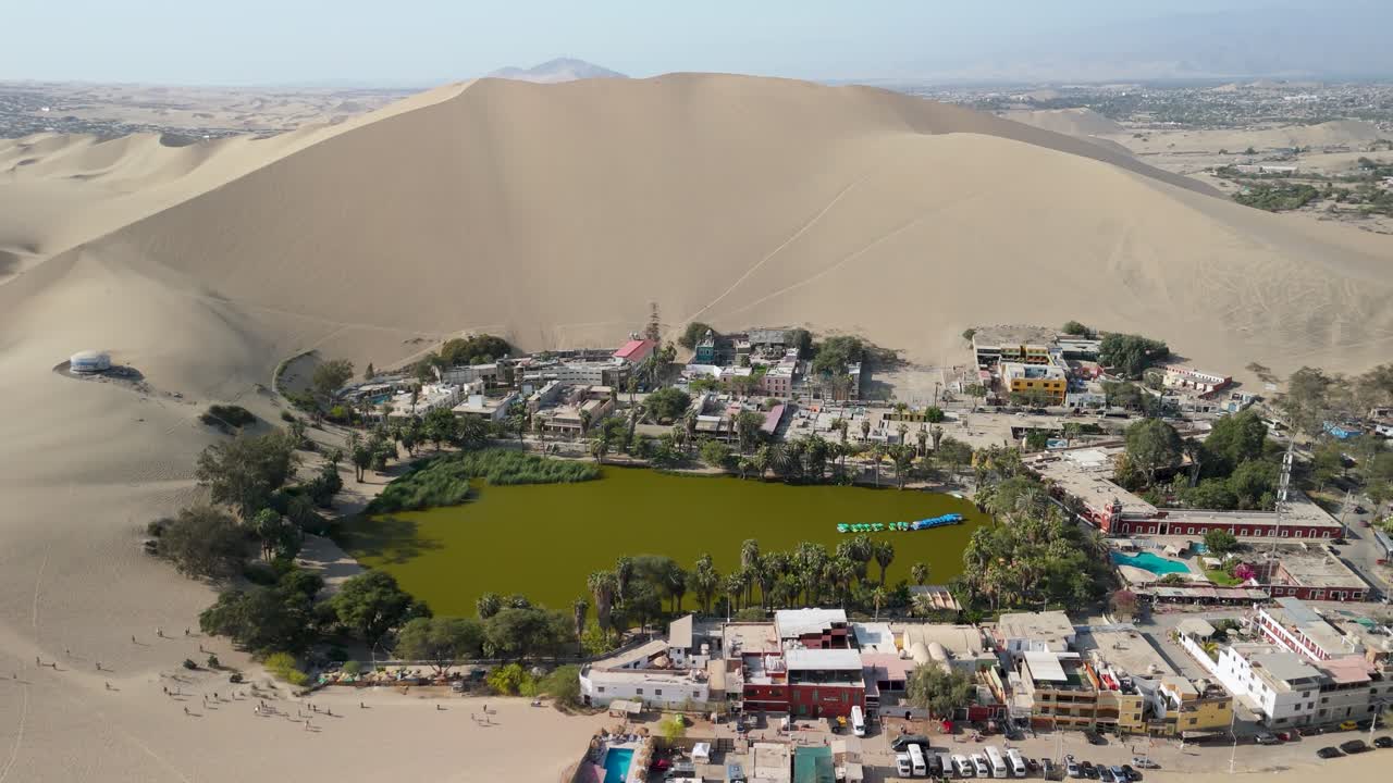 Stunning aerial reveal shot of the Huacachina village and lagoon a lush desert oasis surrounded by massive sand dunes in Ica Peru