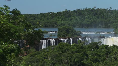 distant waterfalls over tall wild jungle trees, big bright green dense treelife surrounding large body of water pouring down steep ciff edge, wide long waterfall in iguazu falls, brazil