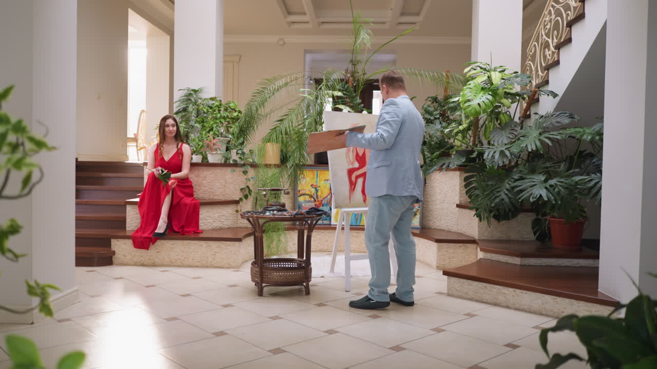 художник рисует соблазнительную женщину в особняке. стильная дама в красном платье сидит среди комнатных растений, в то время как мастер работает на холсте. картина настоящей красоты