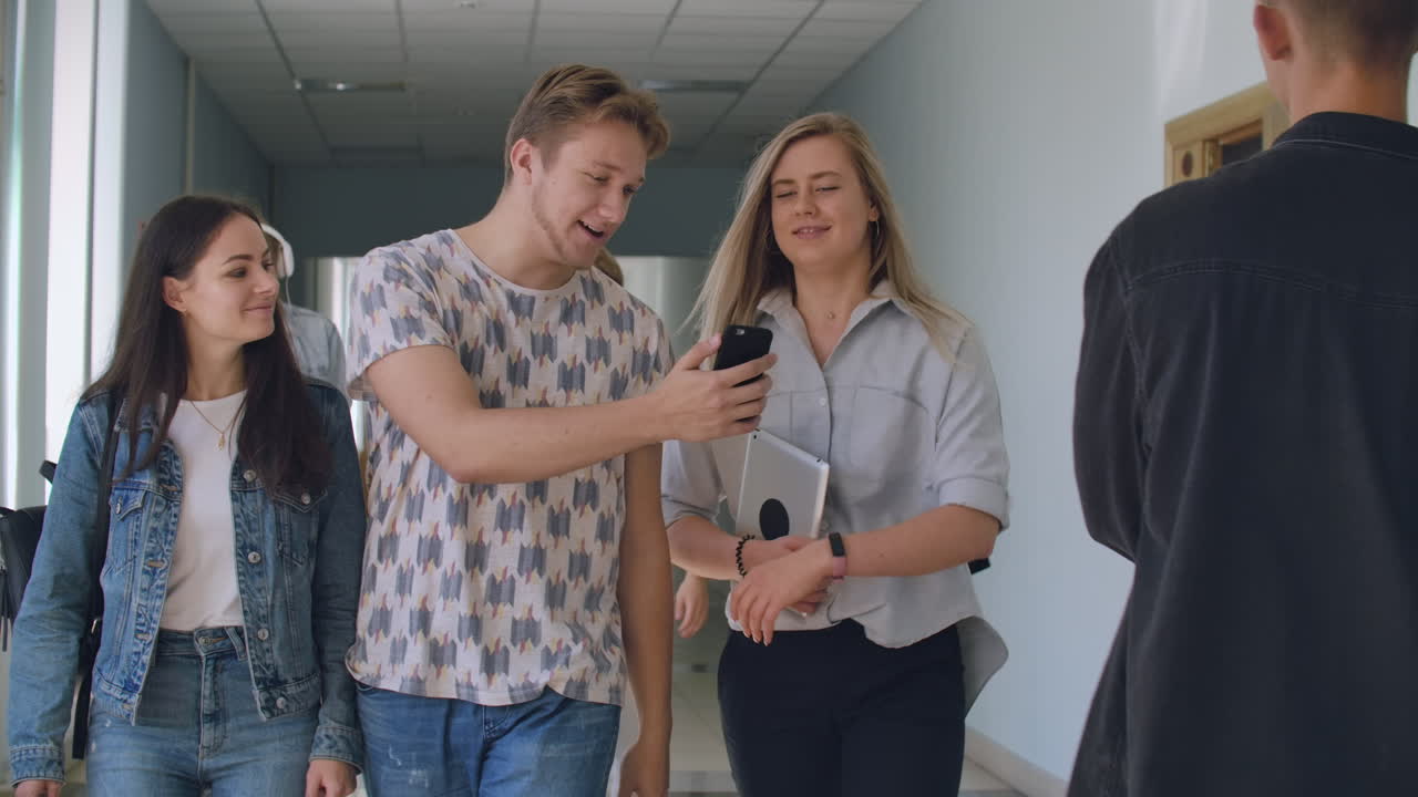 tres estudiantes caminan por el pasillo de la universidad y miran la pantalla del teléfono inteligente y se ríen.