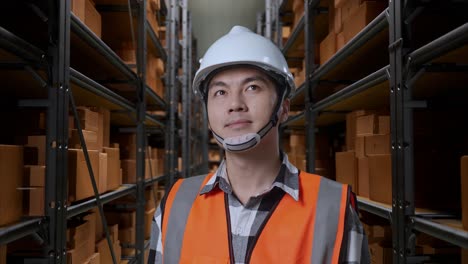 close up of asian male engineer with safety helmet standing in the warehouse with shelves full of delivery goods. looking around, checking the stock on racks