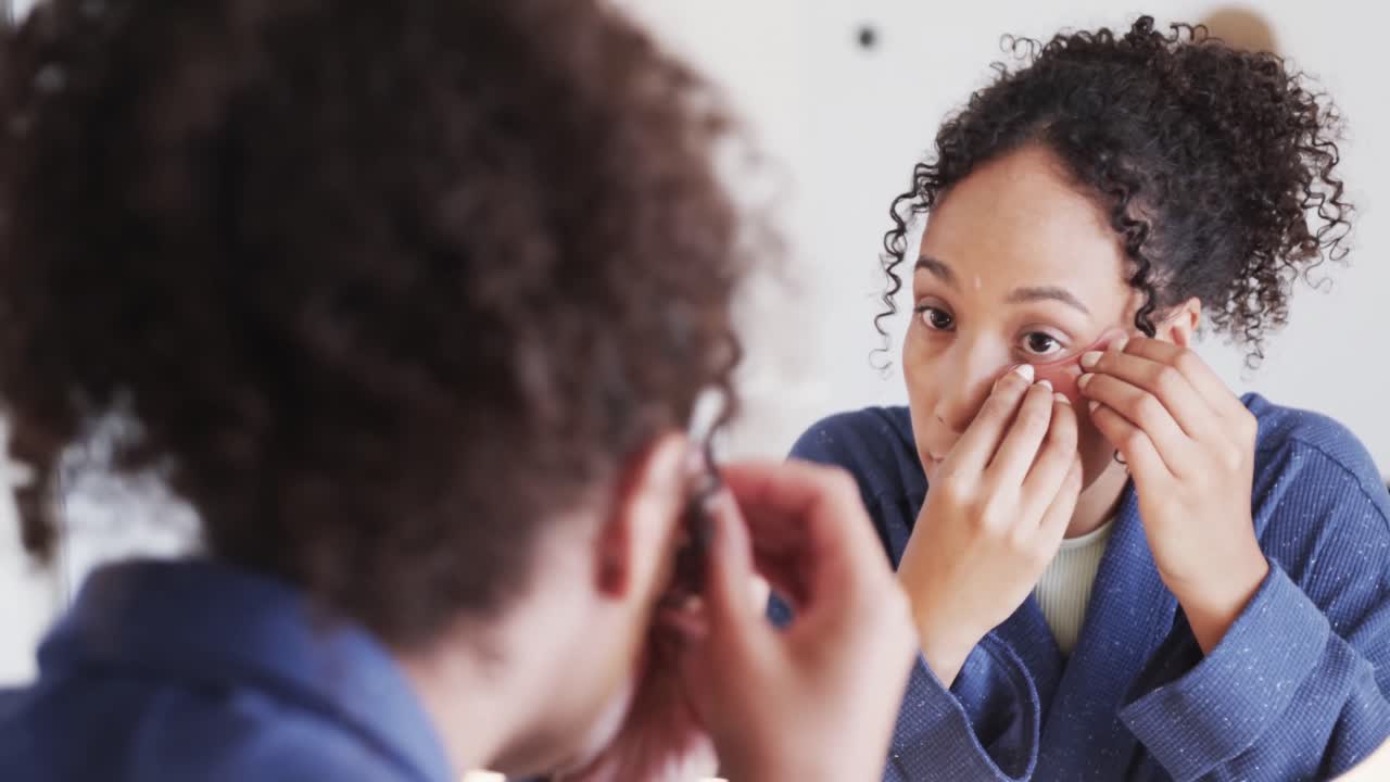 Donna birazziale concentrata che si applica maschere contorno occhi guardandosi allo specchio in un bagno soleggiato, al rallentatore.