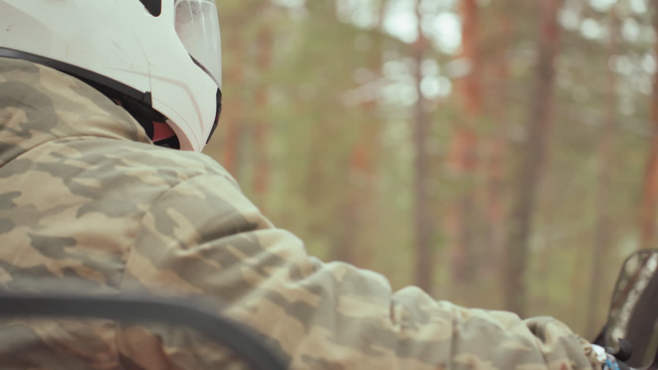 Un pilota camuffato su un ATV nel bosco, con casco bianco e giacca mimetica, manovra tra i pini. Primi piani di profilo e inquadrature posteriori mostrano le mani sul manubrio e il supporto per la action camera, sotto una luce naturale soffusa.