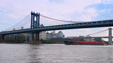a barge moving slowly by the east river under the manhattan bridge. new york skyline at backdrop