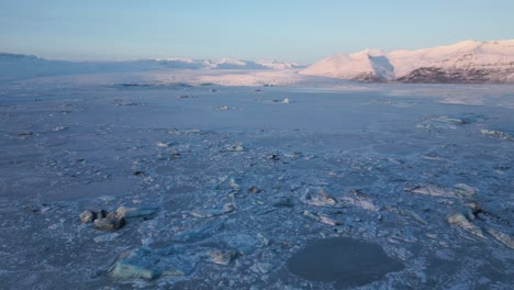 Flying-above-frozen-glacial-lake-towards-mountains-illuminated-by-sunset