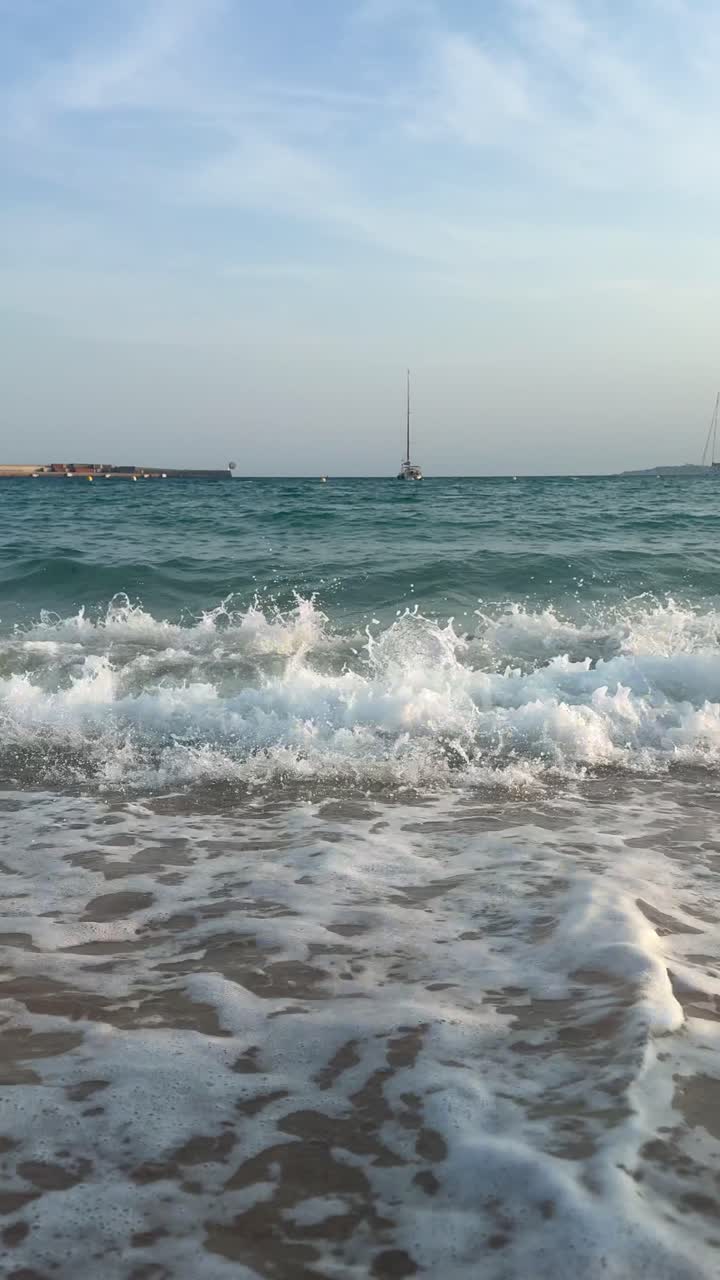 Ocean waves crash on sandy beach with sailboat on horizon, peaceful mood