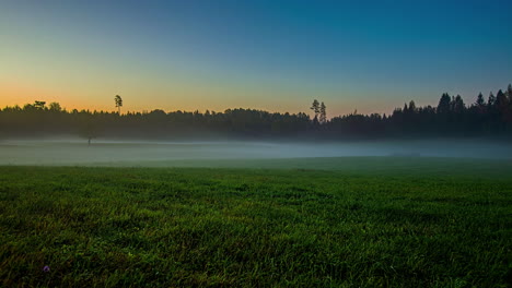 Zeitraffer-Eines-Mit-Tau-Bedeckten-Grasfeldes-Bei-Sonnenaufgang-Mit-Fließendem-Nebel-Im-Hintergrund-Und-Blauem-Und-Orangefarbenem-Himmel