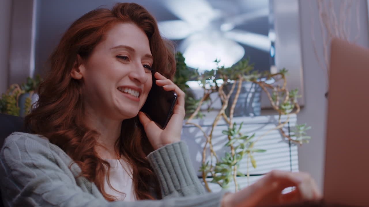 젊은 백인 여성 이 노트북 에서 집 에서 일 하고 있으며, 전화 로 좋은 소식 을 듣고 있다