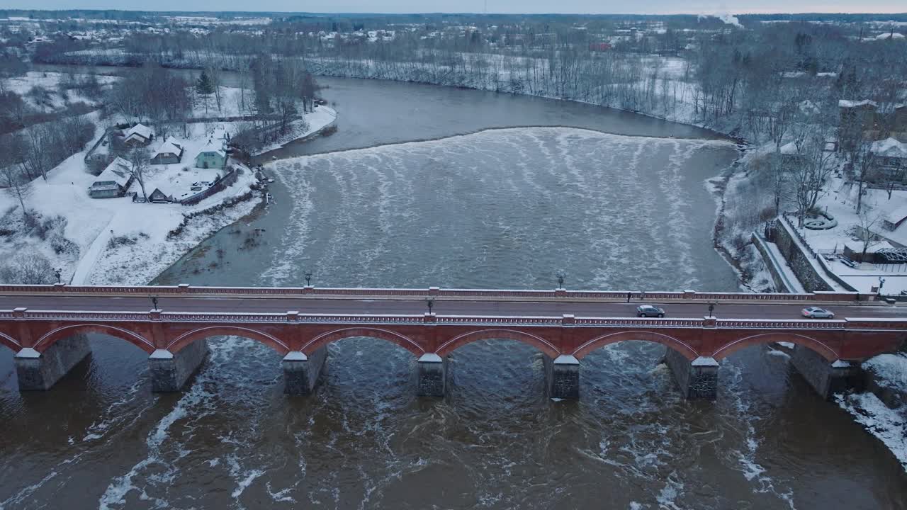 vista aérea de los rápidos del río venta durante la inundación de invierno, viejo puente de ladrillo rojo, kuldiga, letonia, día de invierno nublado, amplia toma de avión no tripulado de ojo de pájaro avanzando
