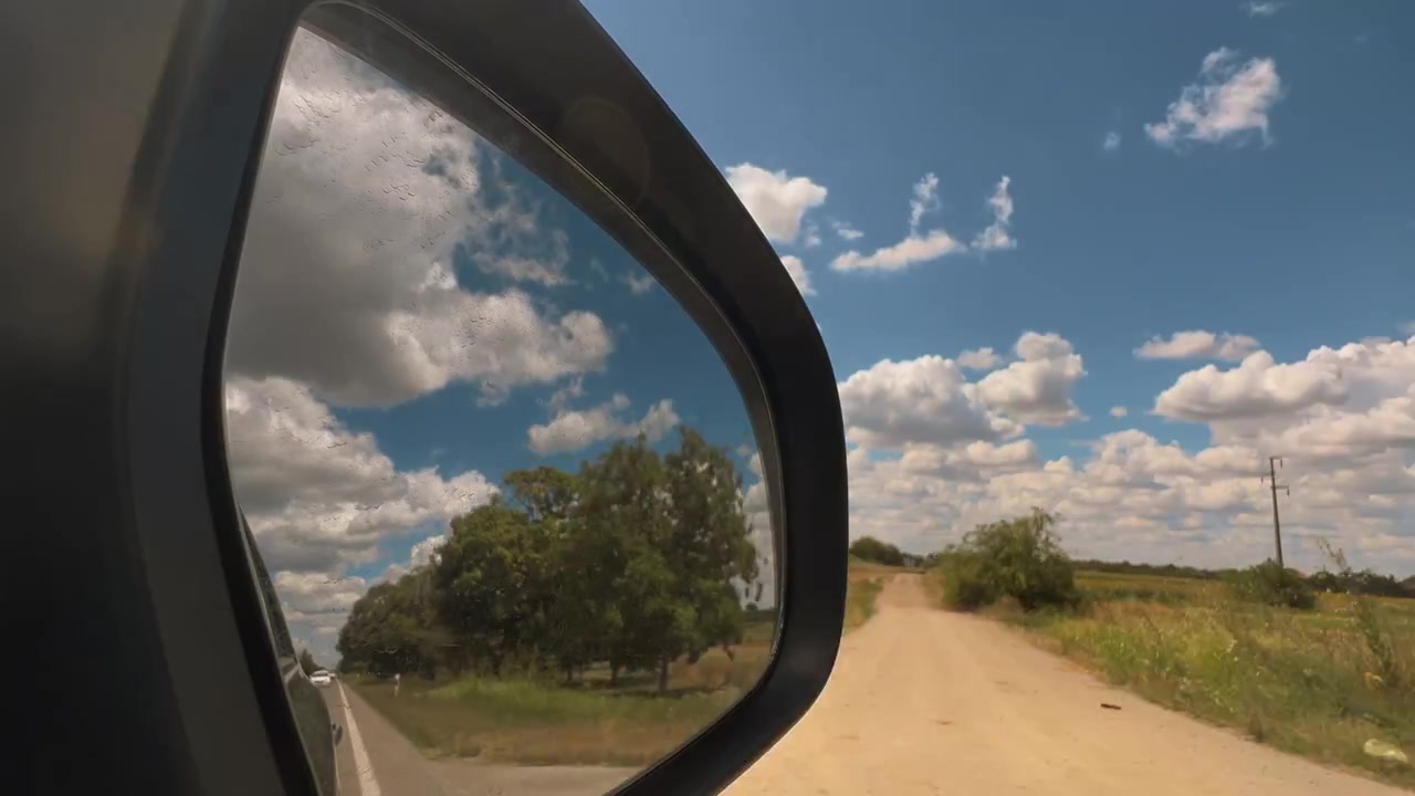 Car side mirror reflecting scenic farmland, roadside trees and a bright summer sky while driving through the countryside. Travel, adventure, transportation and peaceful rural landscape theme. 4K.