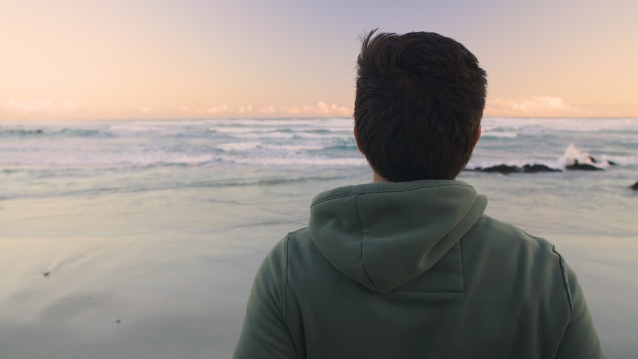 Camera orbits around young man in jogging hoody as he turns and looks at ocean before late afternoon run, as the sun sets over the horizon