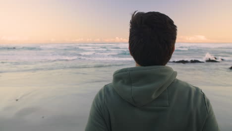 camera orbits around young man in jogging hoody as he turns and looks at ocean before late afternoon run, as the sun sets over the horizon