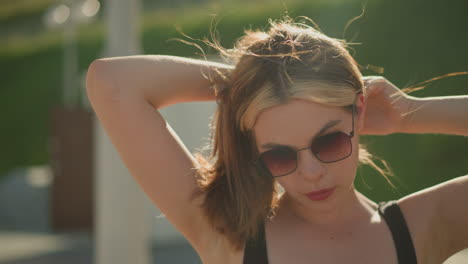 lady adjusts her hair while wearing sunglasses as the wind blows through her hair, the sun radiates on her, creating a warm glow, the background features blurred greenery and poles