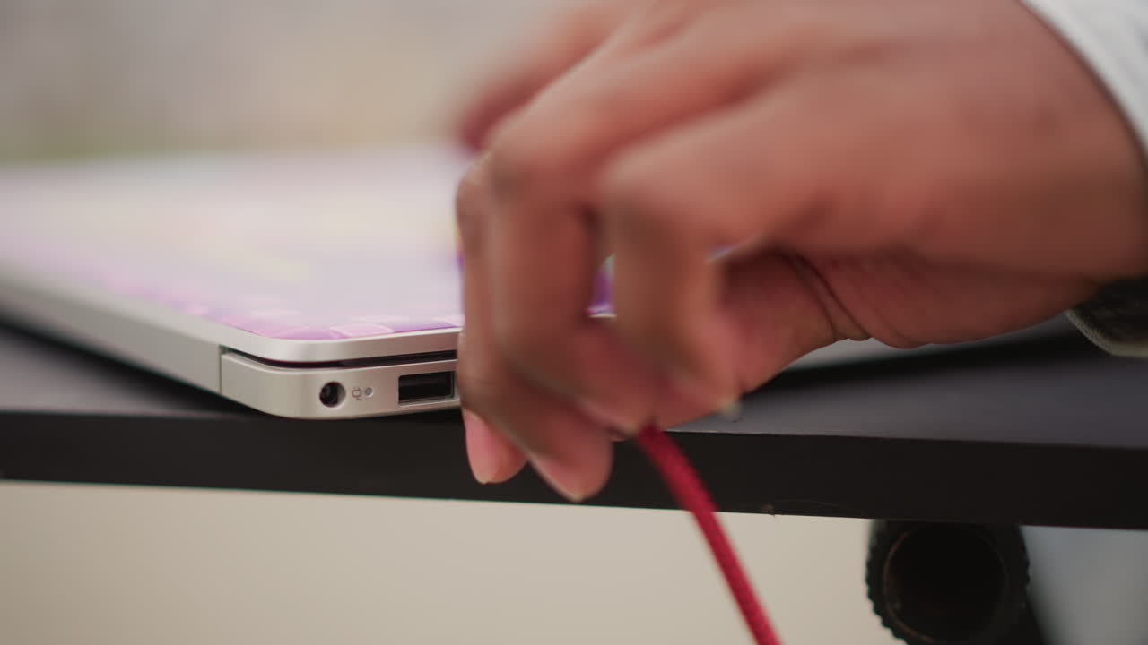 black DJ hands plug laptop cable, closeup of red braided cable inserted into laptop port, soft bokeh background, fingertips adjusting digital setup, software playlist visible, producer preparing live