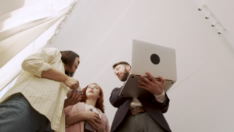 below shot of a man holding a laptop computer and showing something to his female colleagues while standing in the office and talking together