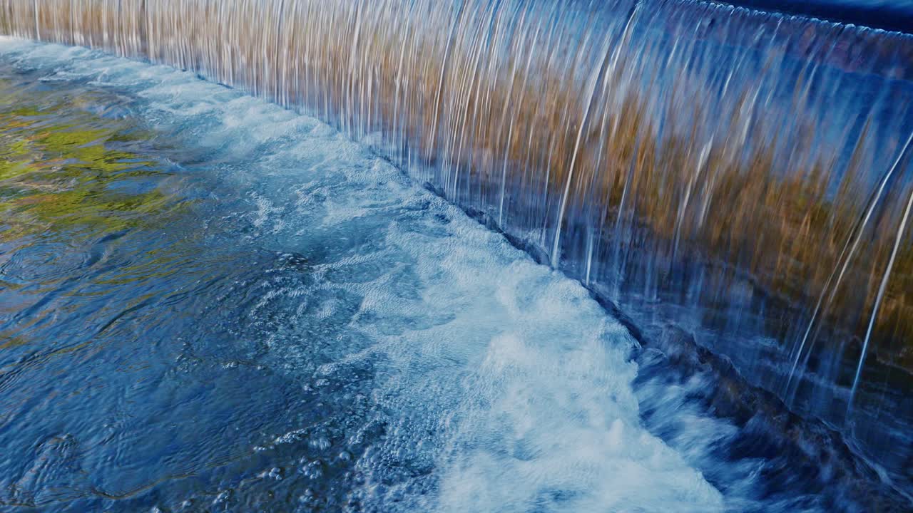 Uno splendido primo piano dell'acqua che scende a cascata su uno sbarramento di pietra curvo in un giardino giapponese. L'acqua forma una cortina liscia, simile a vetro, prima di gettarsi nello stagno increspato sottostante in pieno giorno.