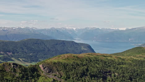 Aerial-shot,-soaring-over-a-tree-lined-ridge,-in-Norway,-revealing-snow-capped-mountains-and-a-Fjord-in-the-background