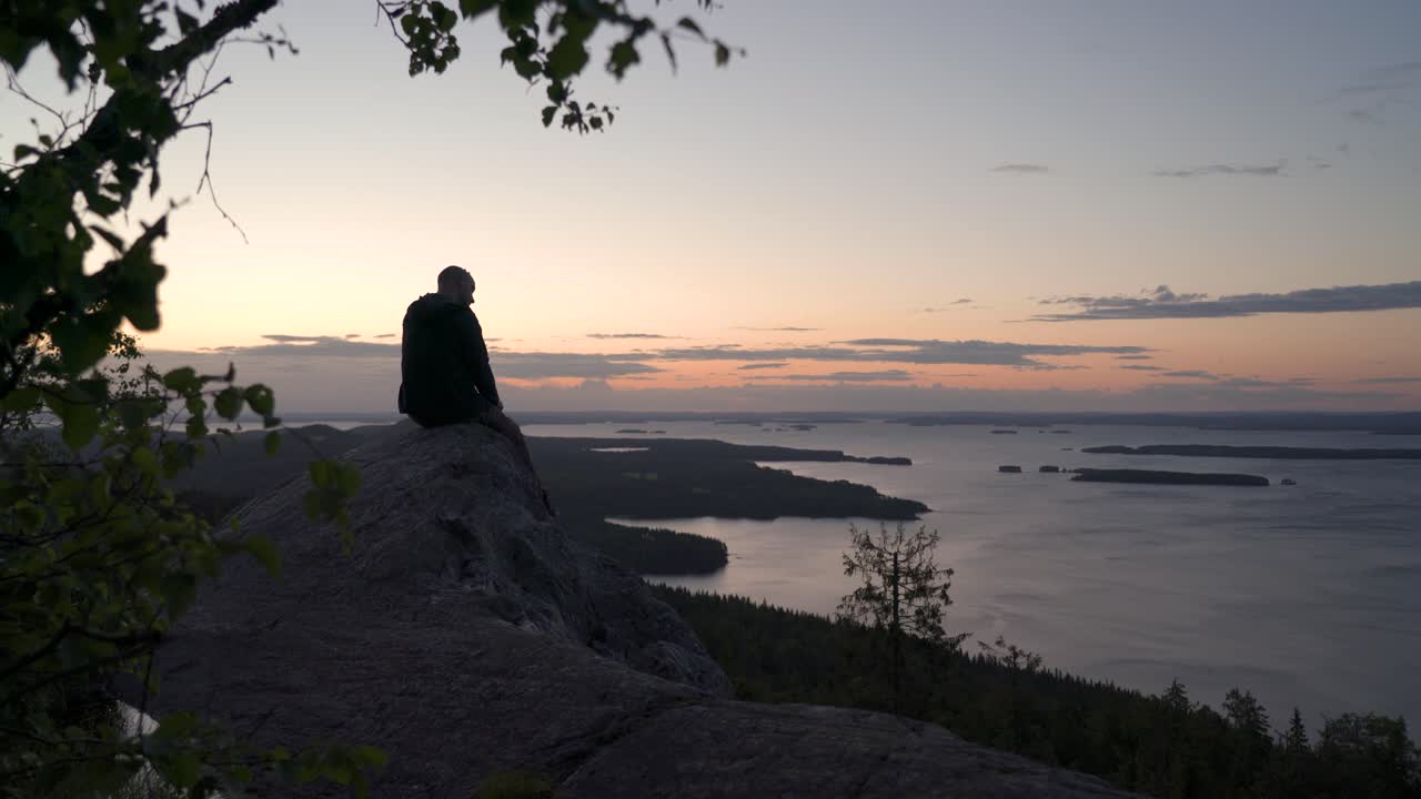 excursionista masculino sentado en un acantilado de montaña nórdico mirando hacia el horizonte y disfrutando de una puesta de sol de verano o un amanecer sobre una gran vista al lago
