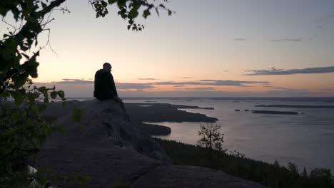 excursionista masculino sentado en un acantilado de montaña nórdico mirando hacia el horizonte y disfrutando de una puesta de sol de verano o un amanecer sobre una gran vista al lago