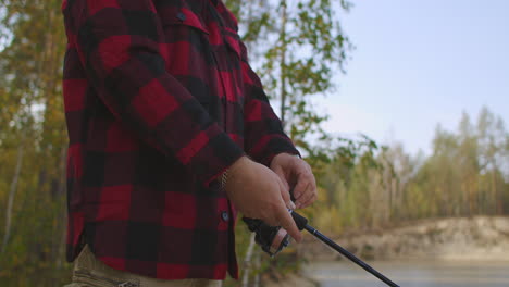 aged man with moustache is fishing on shore of river in forest at autumn day rotating reel of rod and relaxing at nature