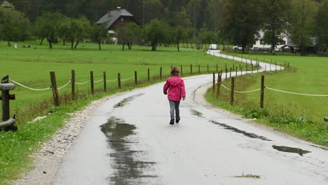 little girl playfully walking in rain on country road, farmhouse in background, alpine valley, hd, slow pan left to right, from behind facing away