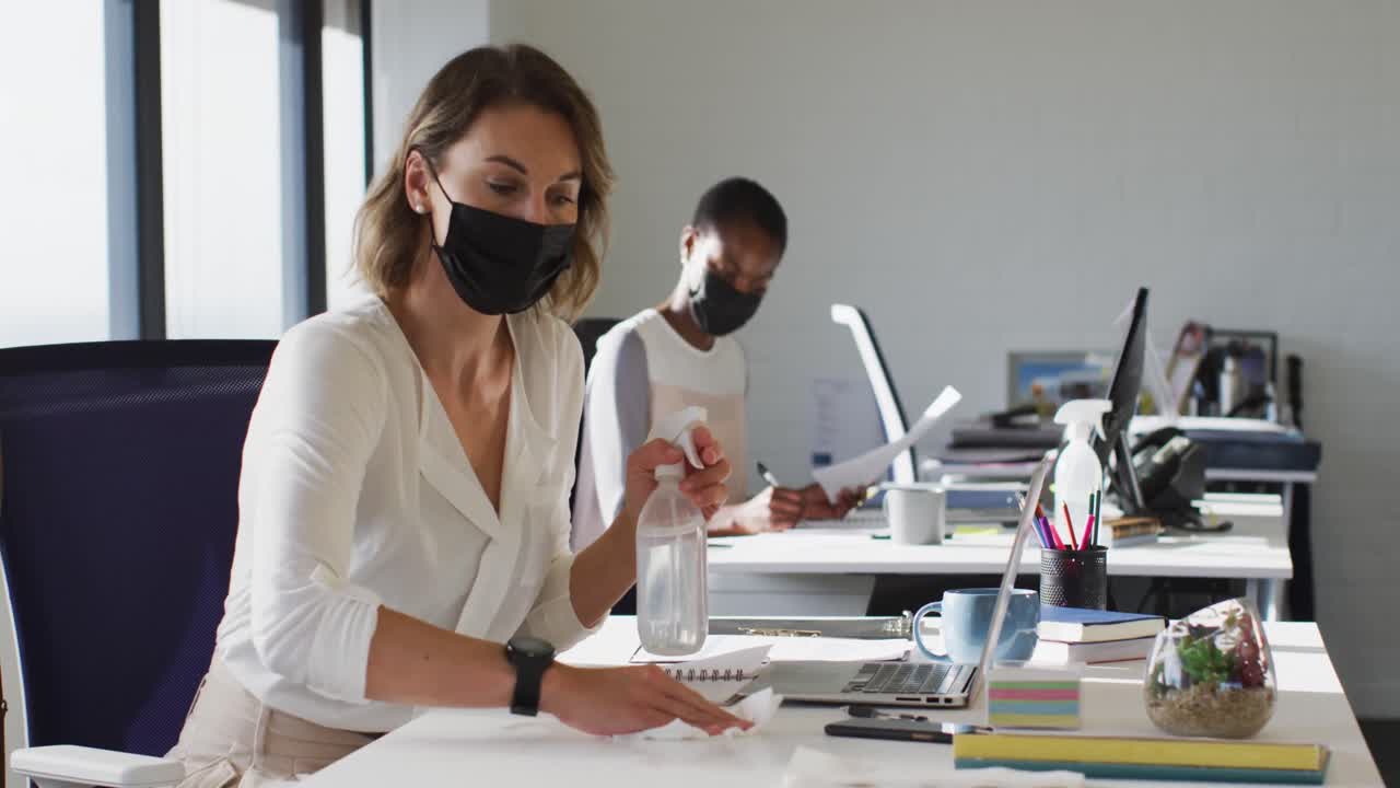 Two diverse female colleagues wearing face mask, sanitizing desk in office