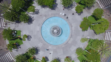 Drone-Circling-Dizengoff-Square,-Tel-Aviv-on-a-hot-summer-day-as-people-sit-and-enjoy-the-shade-of-the-trees-and-a-cyclist-crosses-the-square