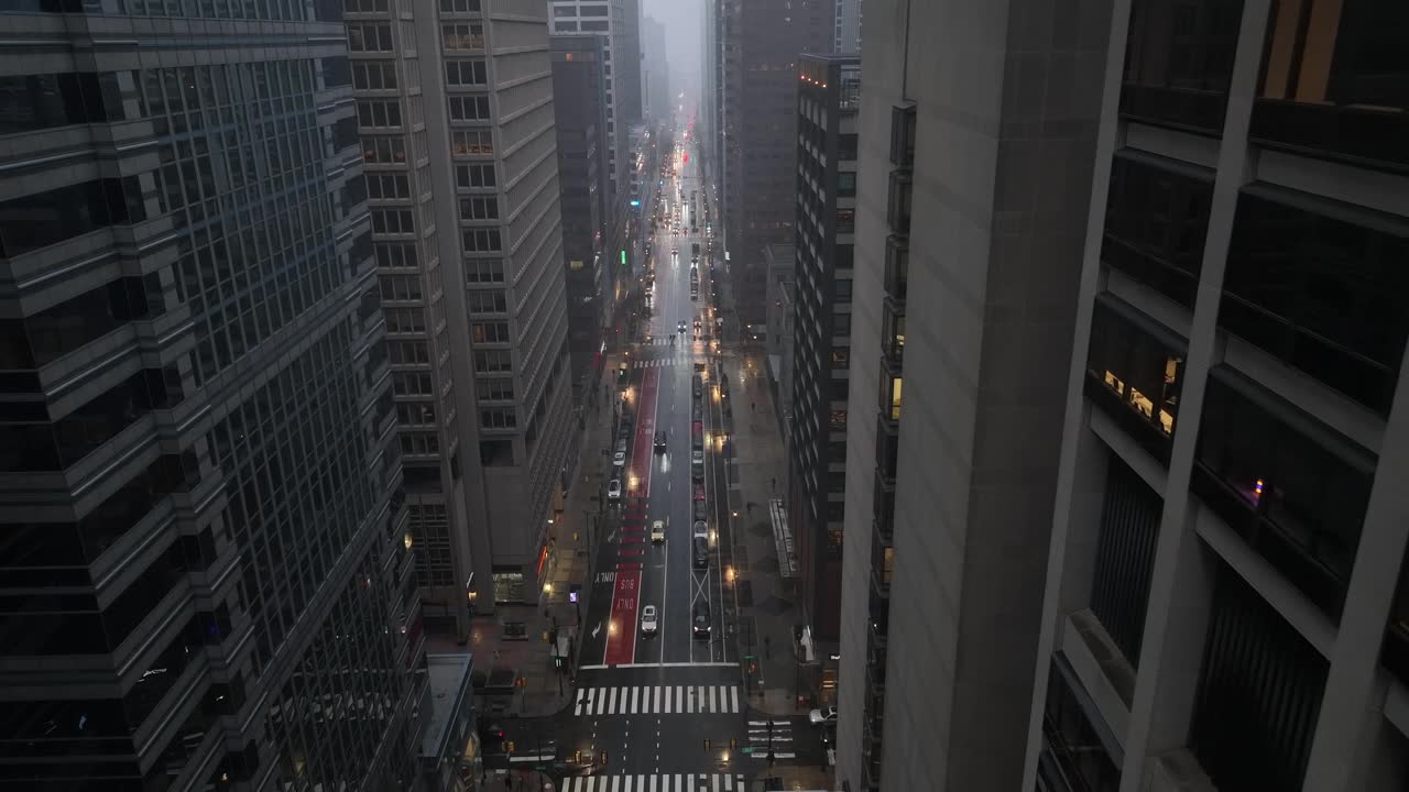 Vista aérea del tráfico urbano durante una nevada en una ciudad estadounidense, con calles mojadas que reflejan las luces, coches circulando, edificios de oficinas de gran altura y una atmósfera invernal gris. Toma descendente.