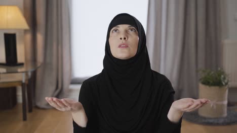 camera moving away from beautiful muslim woman sitting with raised hands, looking up and praying. portrait of young lady in traditional hijab at home. religion, traditional culture.