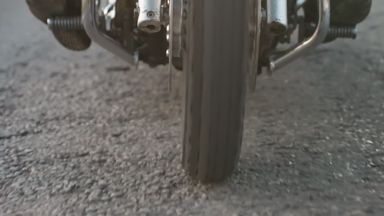 Primo piano dal basso di un motociclista su strada asfaltata con la luce del sole che crea un alone suggestivo intorno al pneumatico, mostrando dettagli cromati, pavimentazione текстурированная e moto dinamica di un veicolo potente