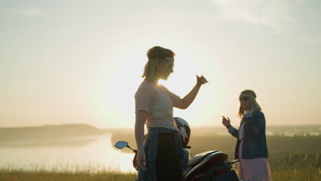 two women, wearing headbands and sunglasses, are joyfully dancing and raising their hands next to a motorcycle in an open grassy field at sunset