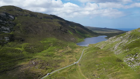 Two-vans-driving-along-a-windy-road-in-Donegal,-overlooking-a-lake-and-valley