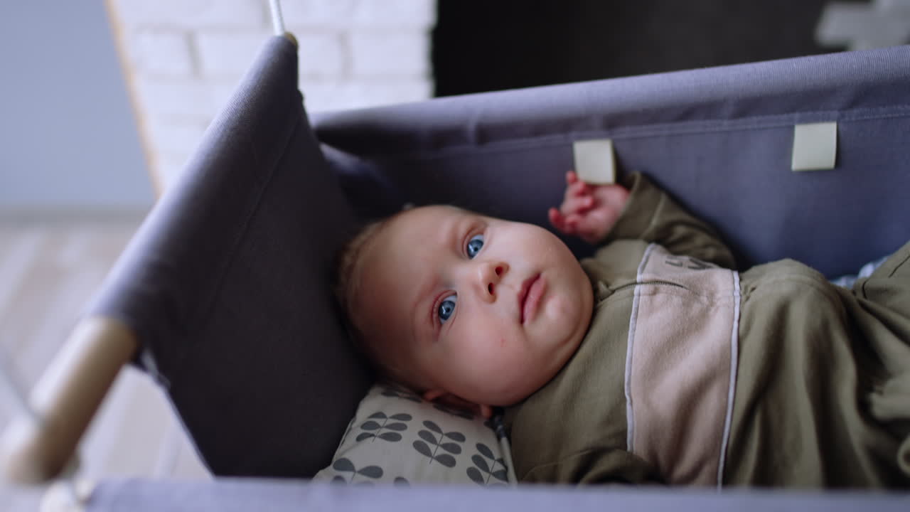 Baby lies in the cradle looking up staying awake. Mom pushes the cradle trying to make kid fall asleep.
