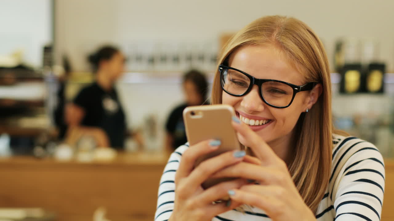 Close-up view of caucasian blonde woman in glasses texting on the phone sitting at a table in a cafe