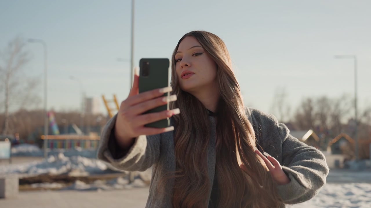 Bela vlogger asiática tirando selfie ao ar livre, mulher segurando o telefone e acenando enquanto fala, cabelo comprido em casaco cinza, cenário urbano de inverno, criação de conteúdo espontâneo e engajamento amigável.