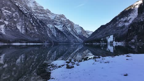 snow covered shoreline with klöntalersee reflecting snowy alps in klöntal, switzerland