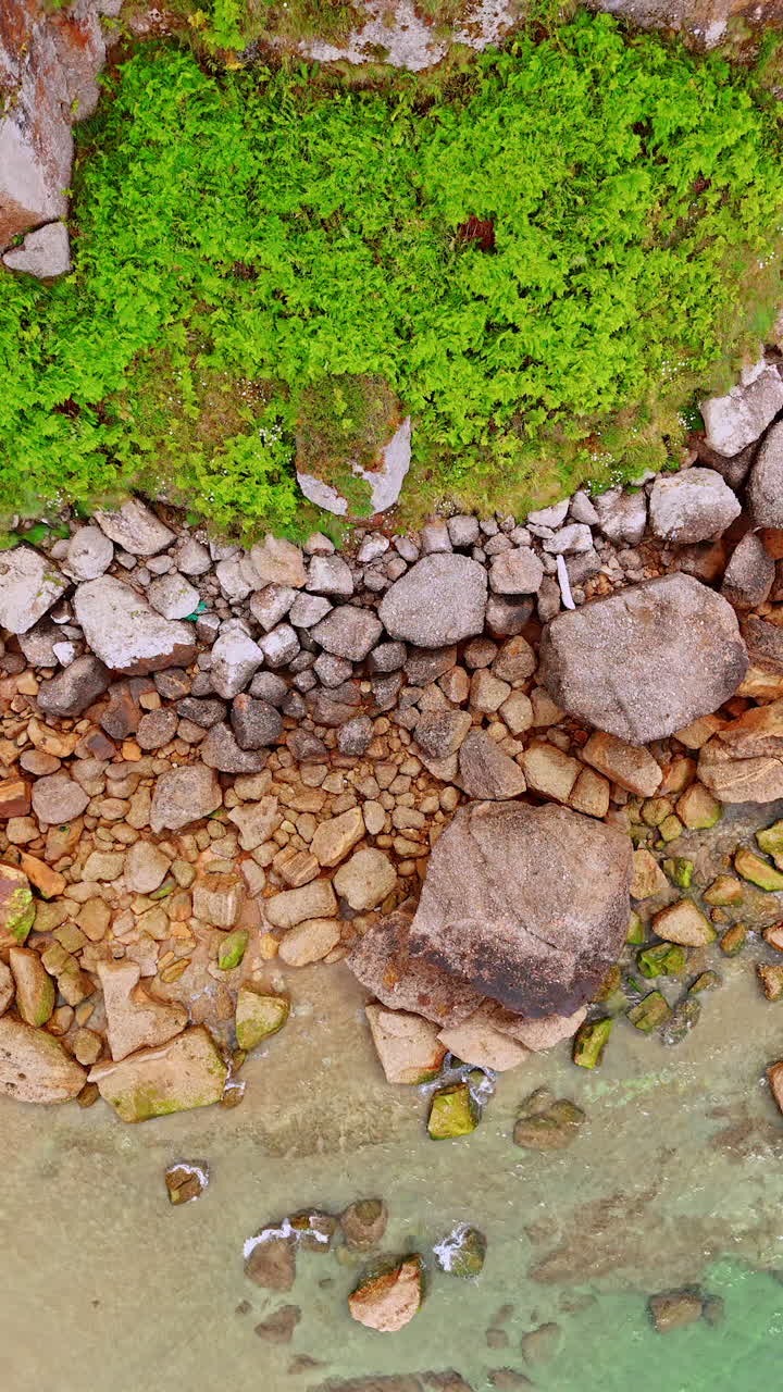 Big and small rocks scattered on the narrow line of a beach. Rising and circling over the shore with clear water. Vertical video.