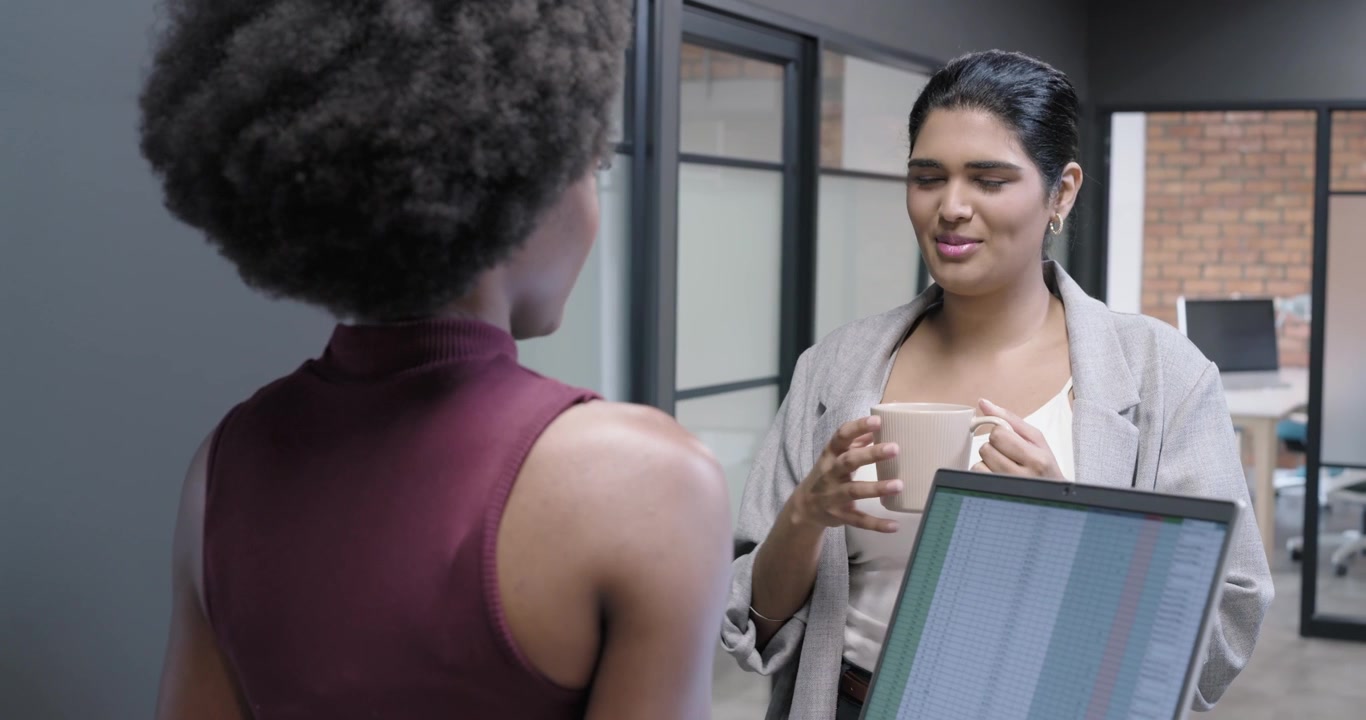 Diverse female coworkers reviewing spreadsheet on laptop at office while listener sipping mug