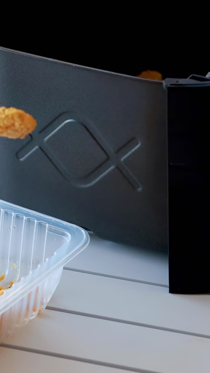A hand uses tongs to transfer breaded food from a plastic tray into a black air fryer basket on a white kitchen surface, under soft natural lighting