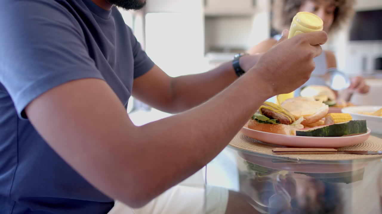 Adding mustard to burger, man enjoying meal with Diverse friends at table