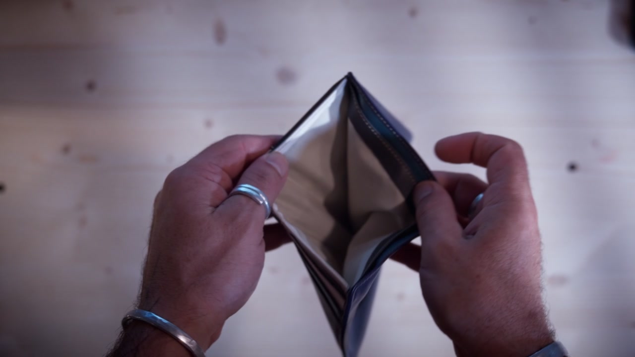 Poverty and financial struggle represented by hands holding an empty black leather wallet. Moody high-contrast visual style in a close-up eye-level view during a stressful moment.