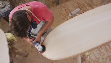 Caucasian-male-surfboard-maker-wearing-a-breathing-face-mask-and-shaping-a-wooden-surfboard-