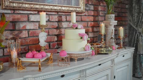 shot of white cake, other desserts and candles on a wooden table with roses in a vase with brick background