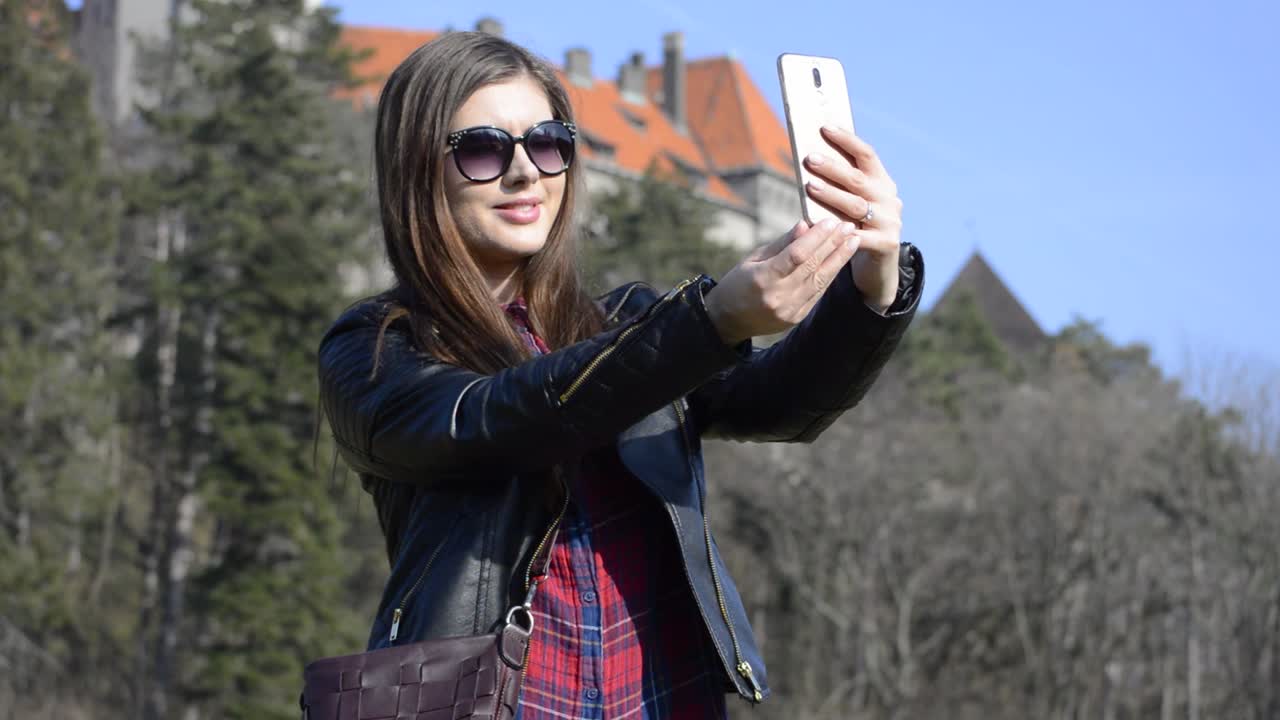 una joven morena con gafas de sol tomando selfies con un castillo en un parque público