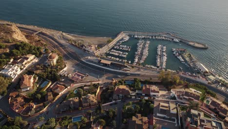 Aerial-flyover-Malaga-City-with-marina-and-Candado-Beach-during-sunset-time-in-Spain---Cars-driving-along-coastal-road